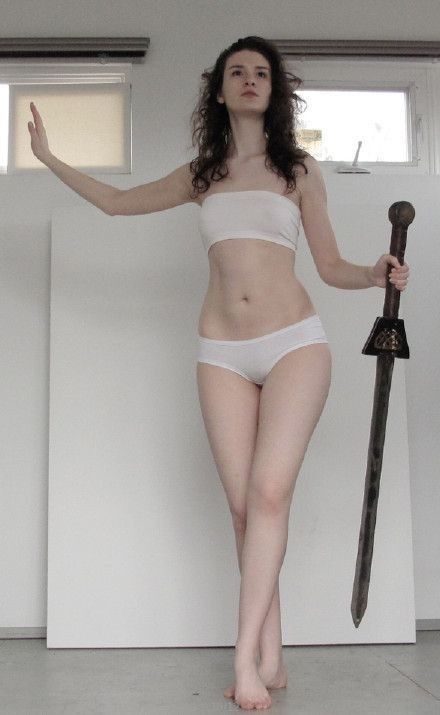 Woman posing confidently with a sword, wearing white attire in a bright room.