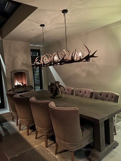 Rustic dining room with antler chandelier, wooden table, tufted chairs, and lit fireplace, creating a cozy atmosphere.