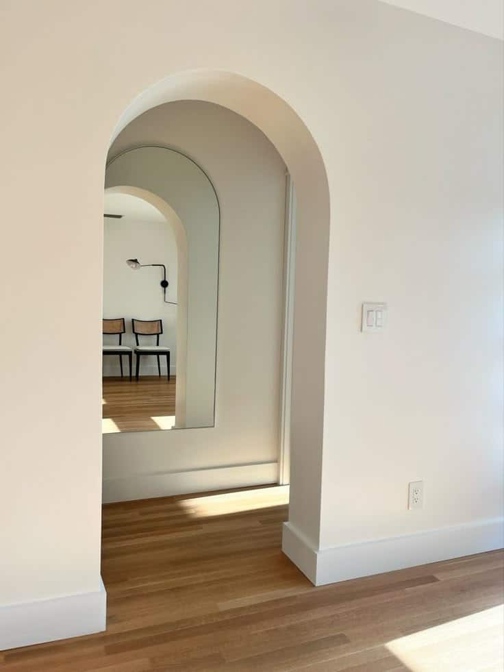 Minimalist white interior with arched doorway, wooden floor, and sunlight casting shadows on the wall.