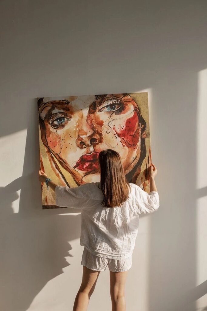 Artistic Inspiration 1 Woman hanging expressive portrait artwork on a wall in soft sunlight. | Sky Rye Design Woman hanging expressive portrait artwork on a wall in soft sunlight.
