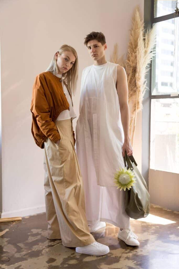 Two individuals in stylish minimalist outfits pose near a window with dried plants in the background.