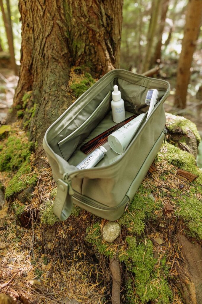 Green toiletry bag with grooming essentials on mossy stump in forest setting.