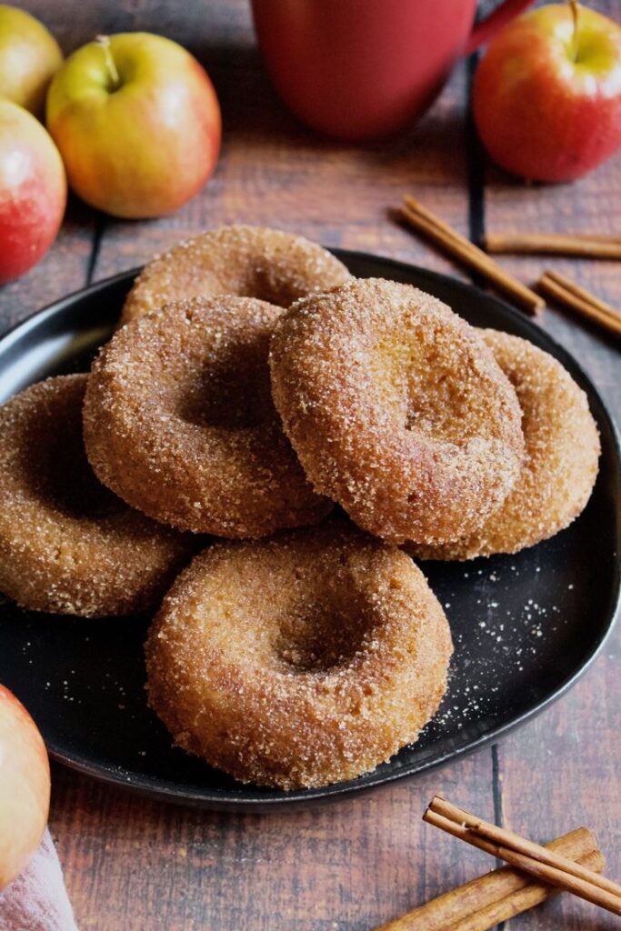 Baked Apple Cider Donuts Plate of apple cider donuts with cinnamon, surrounded by fresh apples and cinnamon sticks on a wooden table. | Sky Rye Design Plate of apple cider donuts with cinnamon, surrounded by fresh apples and cinnamon sticks on a wooden table.