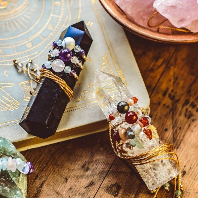 Crystals wrapped with beaded cords on an astrology book, surrounded by gemstones on a wooden table.