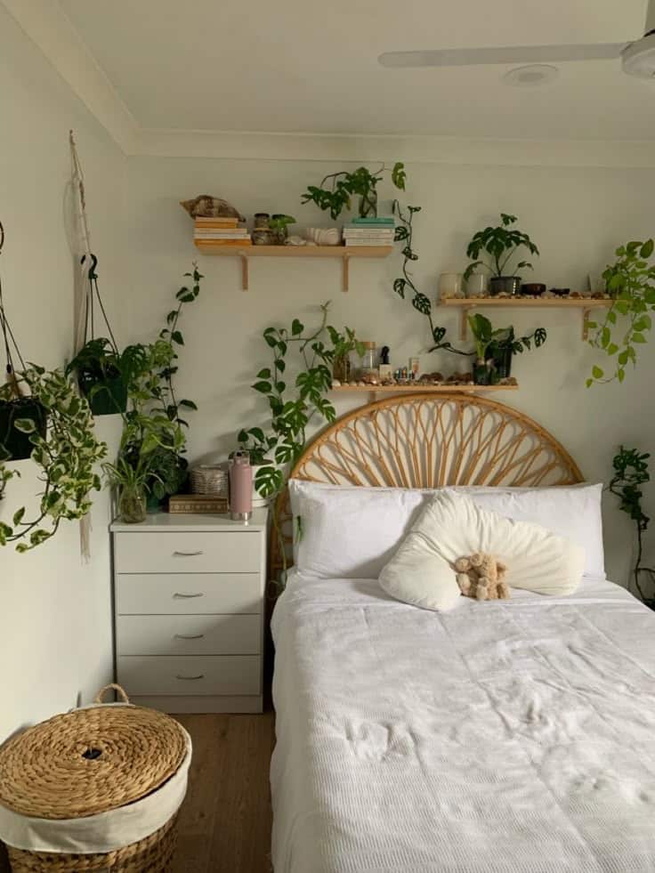 Cozy bedroom with white bedding, wicker headboard, lush green plants, books on shelves, and natural decor elements.
