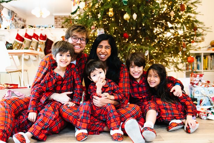 Family in matching red plaid pajamas smiling by a decorated Christmas tree and presents.
