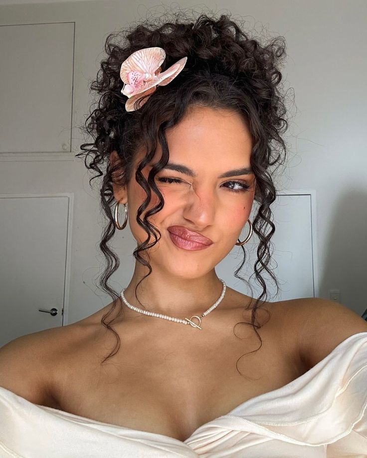Woman winking, wearing a floral hair accessory, off-shoulder dress, pearl necklace, and hoop earrings. Bright smile.