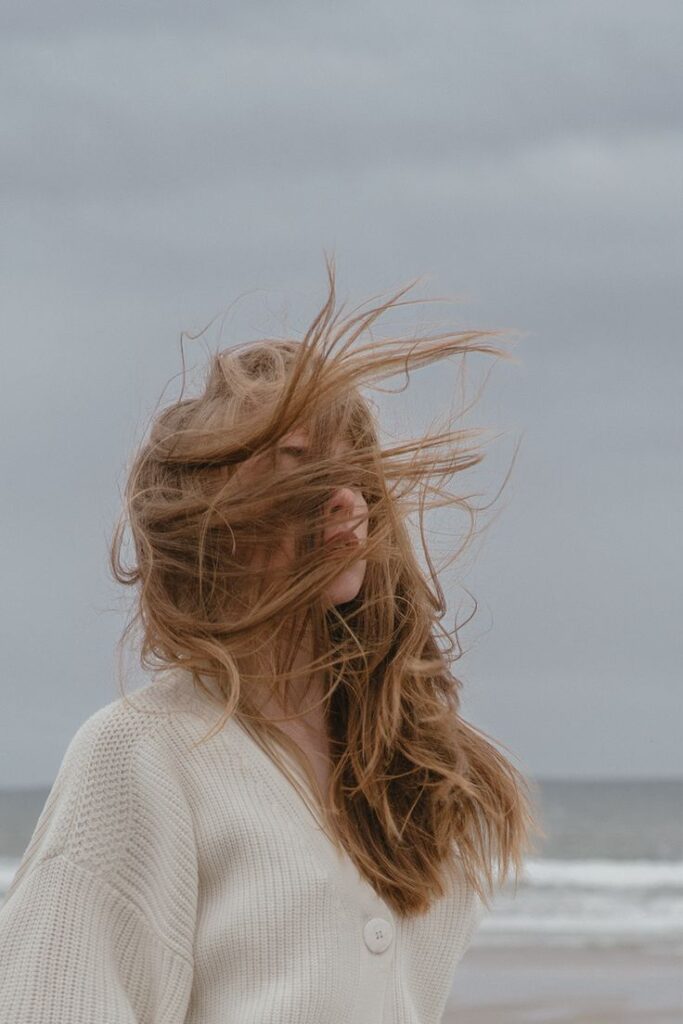 Person with windswept hair stands on a beach under a cloudy sky, wearing a white sweater.