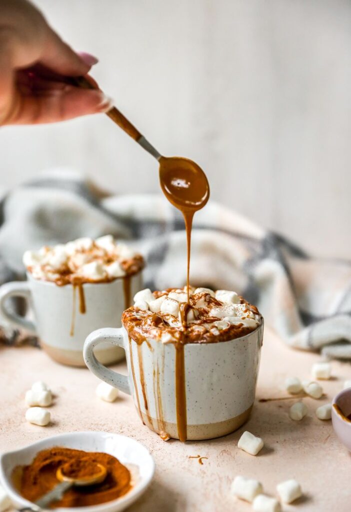 Pouring caramel into hot cocoa with marshmallows in a mug, surrounded by cinnamon for a cozy winter treat.
