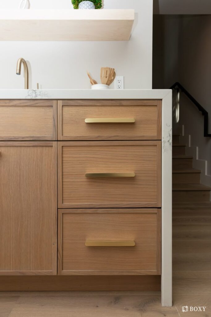 Modern kitchen cabinetry with gold handles, marble counter, and faucet. Shelf with plant accent above.