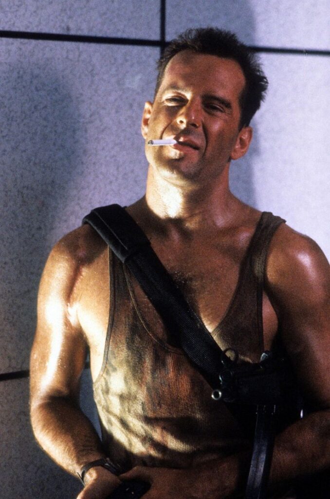 Man in a dirty tank top, holding a cigarette, with a rugged expression and strap over his shoulder, in an intense scene.