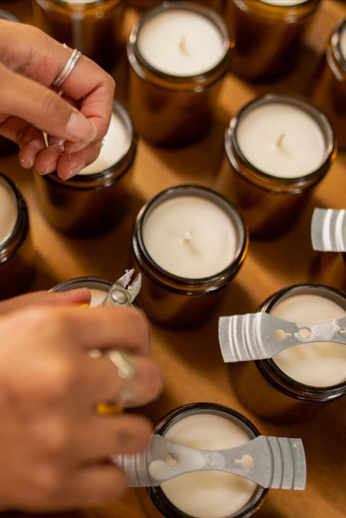 Candle studio Person crafting soy candles in amber jars, adjusting wicks with trimmers in a cozy workspace setting. | Sky Rye Design Person crafting soy candles in amber jars, adjusting wicks with trimmers in a cozy workspace setting.