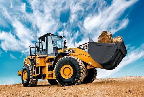 Yellow front loader carrying large rocks on a construction site under a blue sky with clouds.