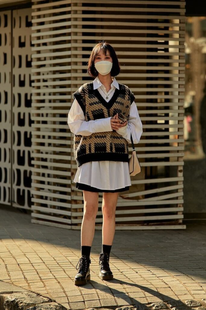 Fashionable woman in plaid sweater vest and mask, standing on city street in sunlight.