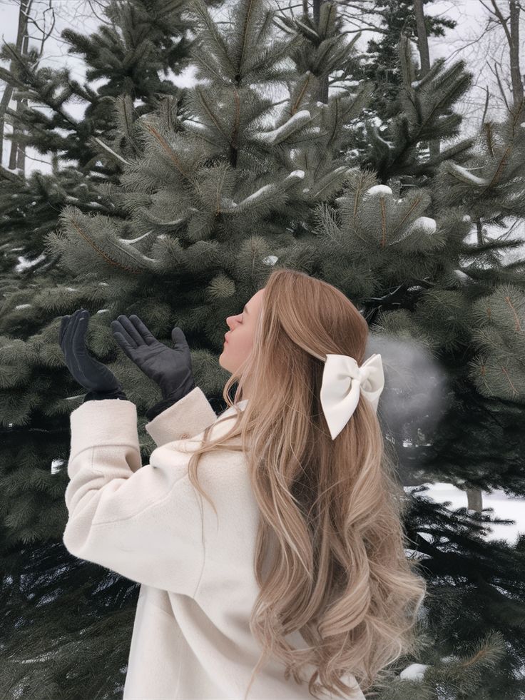Woman in winter coat with bow in hair enjoys snowy day by pine tree.