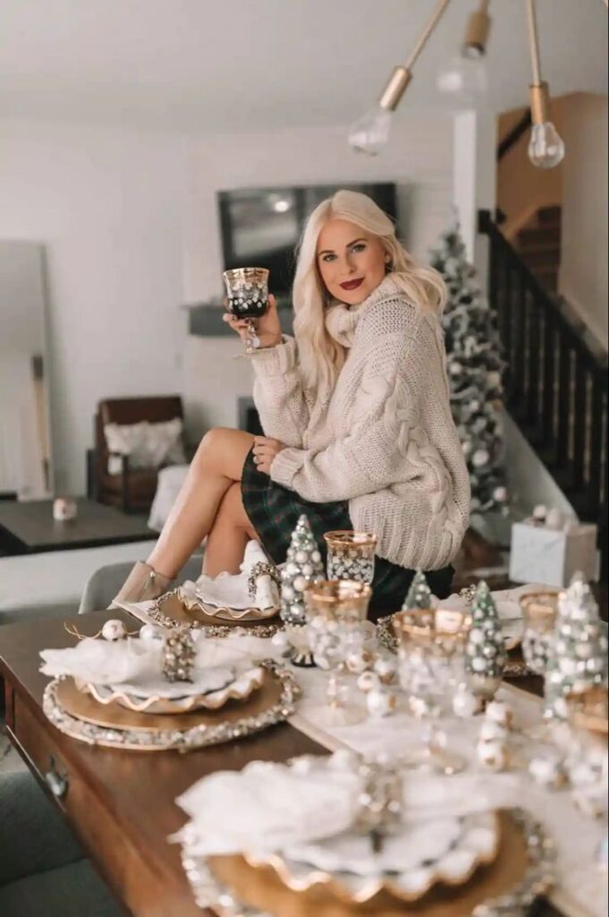 Woman in cozy sweater enjoying holiday drink at festive dining table with Christmas decor and ornaments.