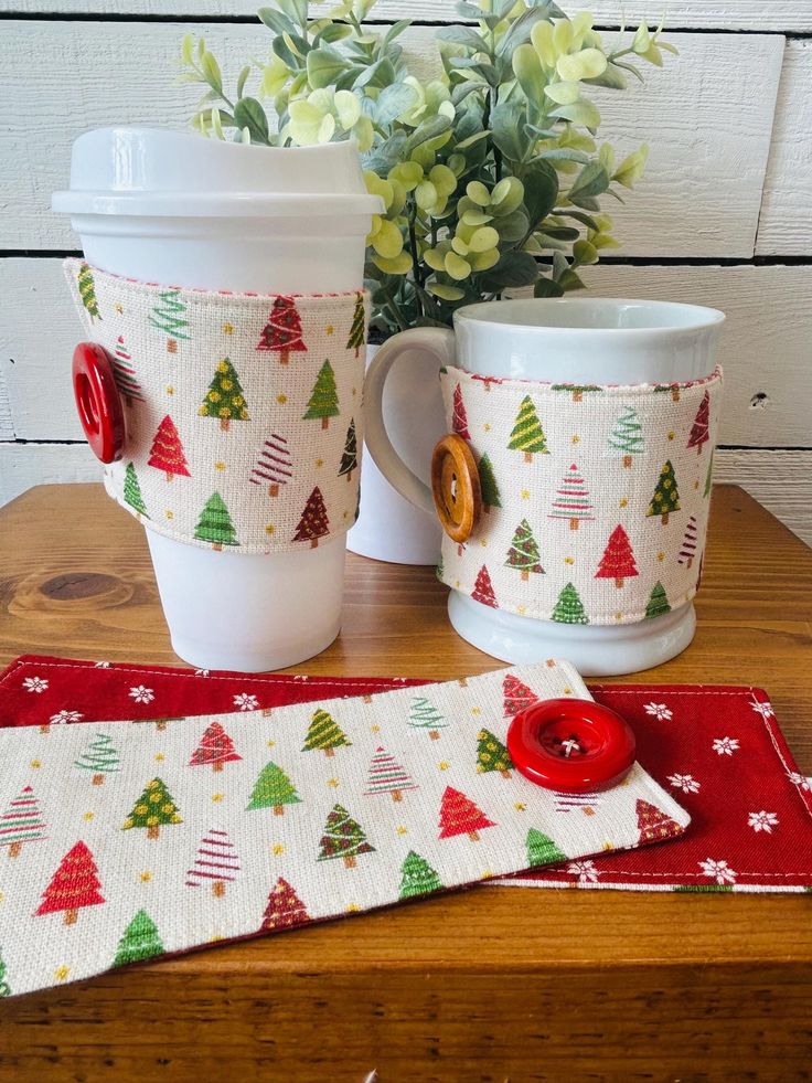 Christmas Tree Reusable Coffee Sleeve_ Holiday Fabric Cozy Festive coffee mugs with holiday tree patterns and cozy fabric wraps, adorned with red buttons, on a wooden table. | Sky Rye Design Festive coffee mugs with holiday tree patterns and cozy fabric wraps, adorned with red buttons, on a wooden table.
