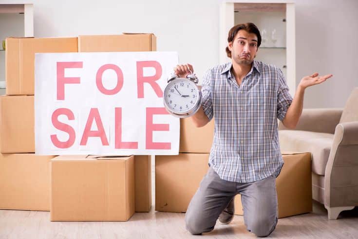 Cities Where the Most Homes Experienced a Price Cut Man kneeling by For Sale sign with boxes, holding clock and shrugging in confusion. | Sky Rye Design Man kneeling by For Sale sign with boxes, holding clock and shrugging in confusion.