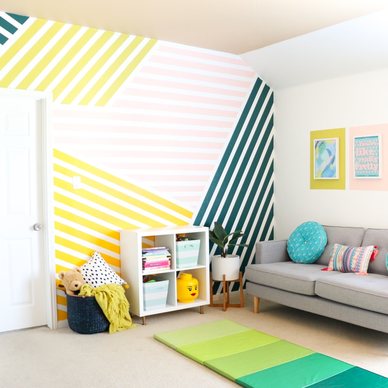 Bright playroom with colorful striped wall, cozy gray sofa, cube storage, and playful decor.