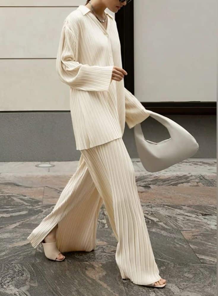 Woman in chic beige pleated outfit with matching handbag and sandals, walking stylishly on textured flooring.