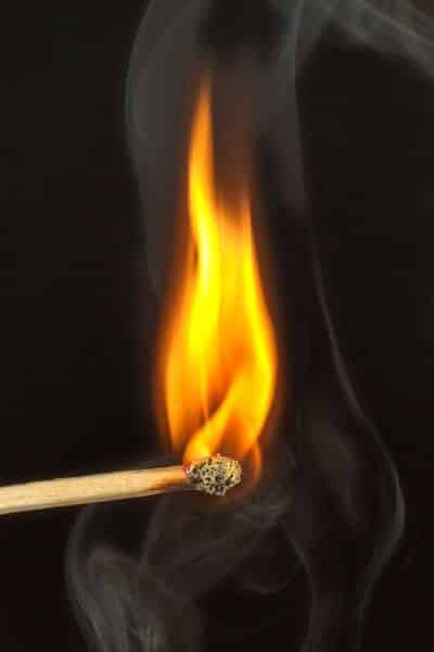Lit matchstick with bright orange flame against a black background, emitting smoke.
