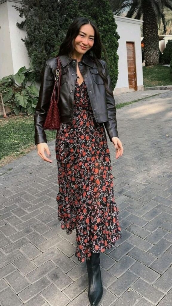 Como usar vestido no inverno Woman in floral dress and leather jacket walking on paved street, carrying red bag, with greenery backdrop. | Sky Rye Design Woman in floral dress and leather jacket walking on paved street, carrying red bag, with greenery backdrop.