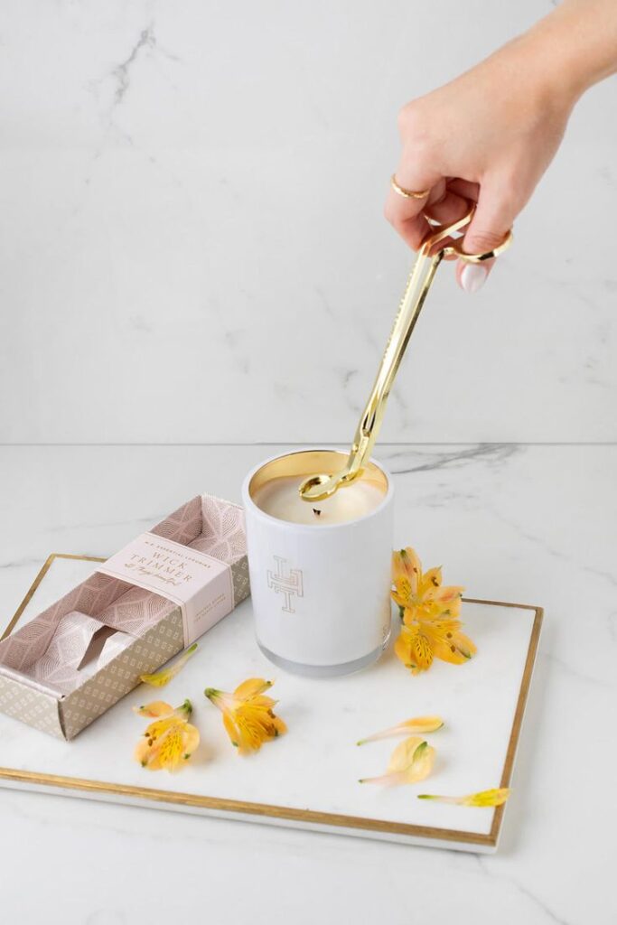 Coupe-Mèche de Luxe Hand using gold wick trimmer on white candle; decorative flowers and trimmer box on marble tray. | Sky Rye Design Hand using gold wick trimmer on white candle; decorative flowers and trimmer box on marble tray.