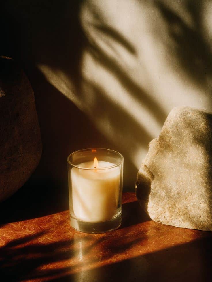 Created by Midjourney prompt ↓ A lit candle in a glass holder on a wooden table, surrounded by rocks, casting warm shadows. Cozy and serene ambiance. | Sky Rye Design A lit candle in a glass holder on a wooden table, surrounded by rocks, casting warm shadows. Cozy and serene ambiance.