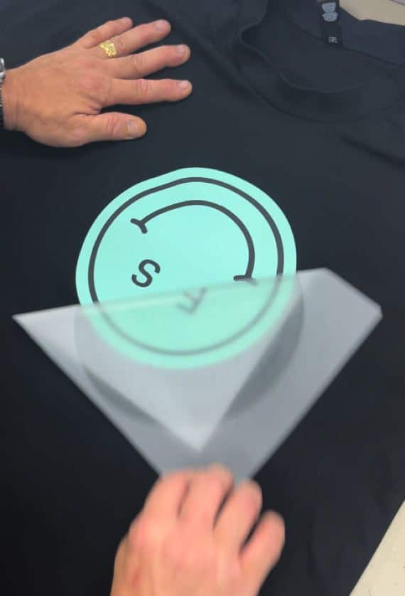 Custom Printed DTF Transfers Sydney Press and Peel Person applying circular teal logo onto black t-shirt with a transparent sheet. | Sky Rye Design Person applying circular teal logo onto black t-shirt with a transparent sheet.