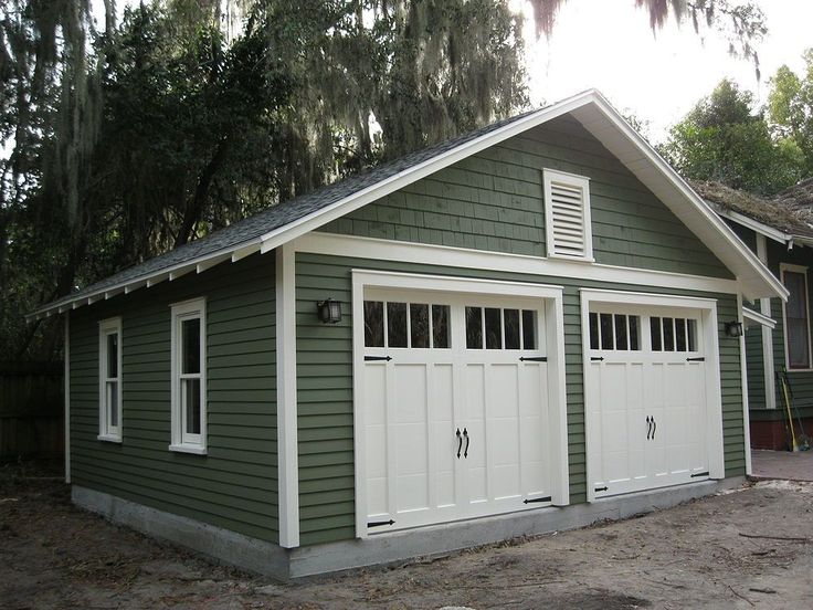 Green and white garage with classic barn-style doors surrounded by trees and moss in a wooded setting.