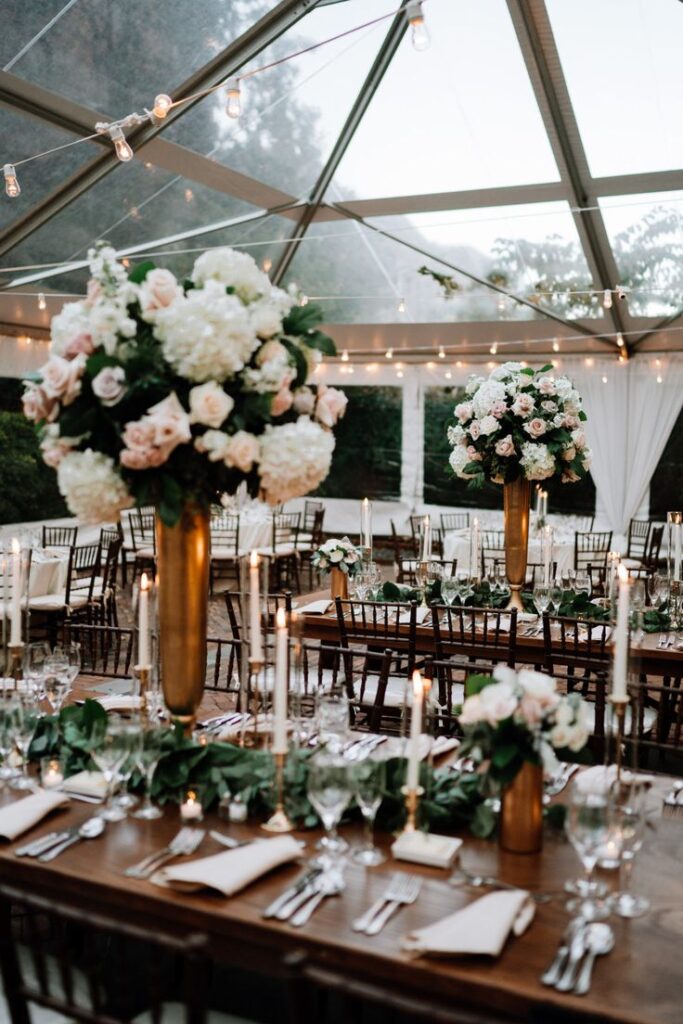 Elegant wedding reception under a glass tent with floral centerpieces and candlelit tables.