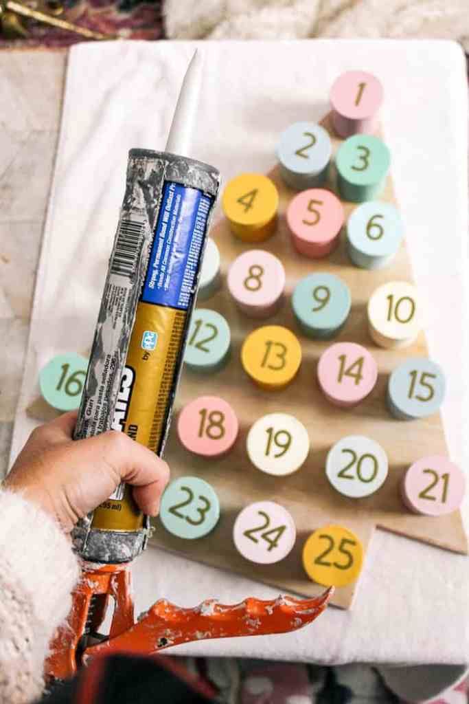 DIY Christmas Advent Calendar Person holding a caulking gun next to a DIY painted block calendar with numbers 1 to 25. | Sky Rye Design Person holding a caulking gun next to a DIY painted block calendar with numbers 1 to 25.