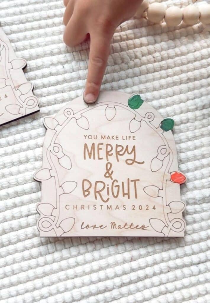 Child pointing at a wooden Christmas card with lights design, saying “Merry & Bright, Christmas 2024, love matters.”