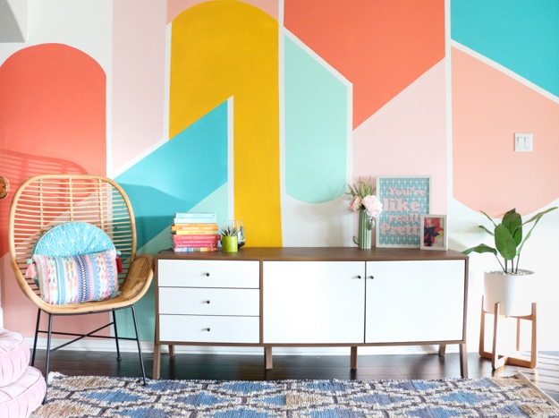 Colorful modern living room with geometric wall, white sideboard, cozy chair, books, and plants.