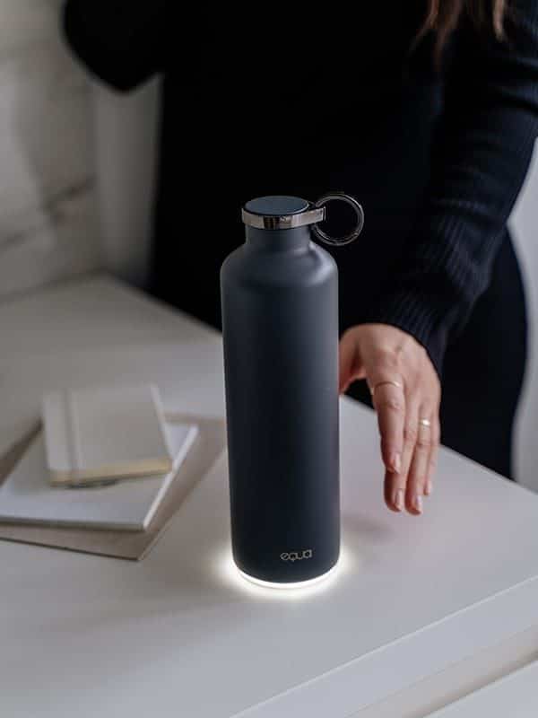 Sleek black water bottle with a glowing base on a desk, beside a person’s hand and notebooks.