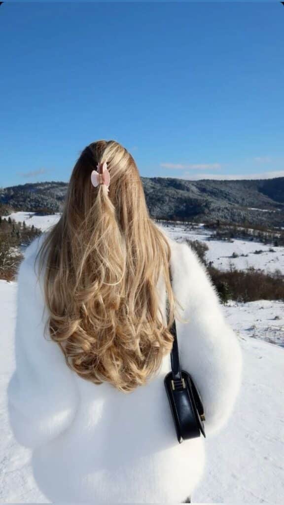 Woman in white coat with long blonde hair and clip, standing in snowy mountain landscape.