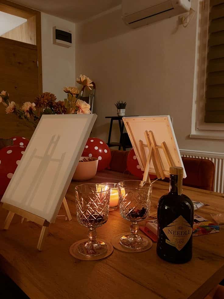 Date night ✨️ Cozy painting setup with easels, candles, wine glasses, and a bottle of gin on a wooden table in a dimly lit room. | Sky Rye Design Cozy painting setup with easels, candles, wine glasses, and a bottle of gin on a wooden table in a dimly lit room.