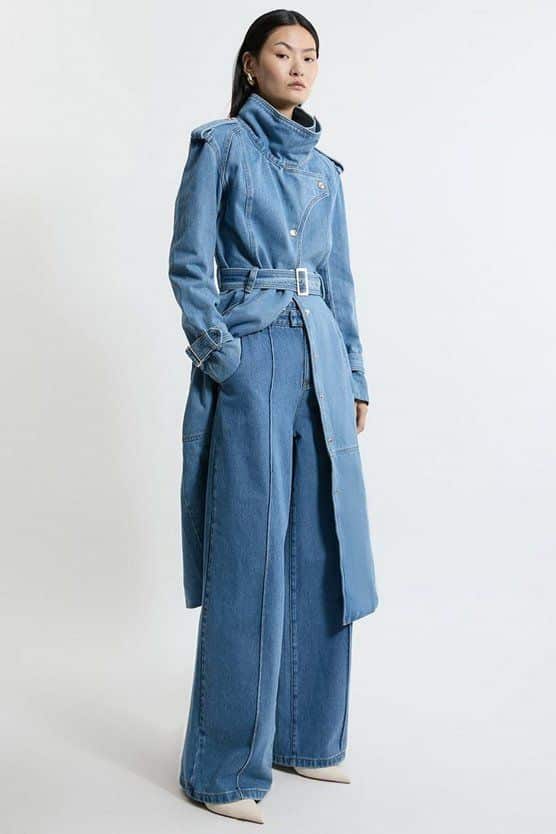 Woman in denim trench coat and wide-leg jeans posing confidently against a plain background.