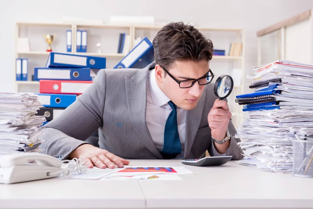 Depositphotos_161191288_XL Businessman examines documents with magnifying glass, surrounded by stacks of paperwork in an office setting. | Sky Rye Design Businessman examines documents with magnifying glass, surrounded by stacks of paperwork in an office setting.