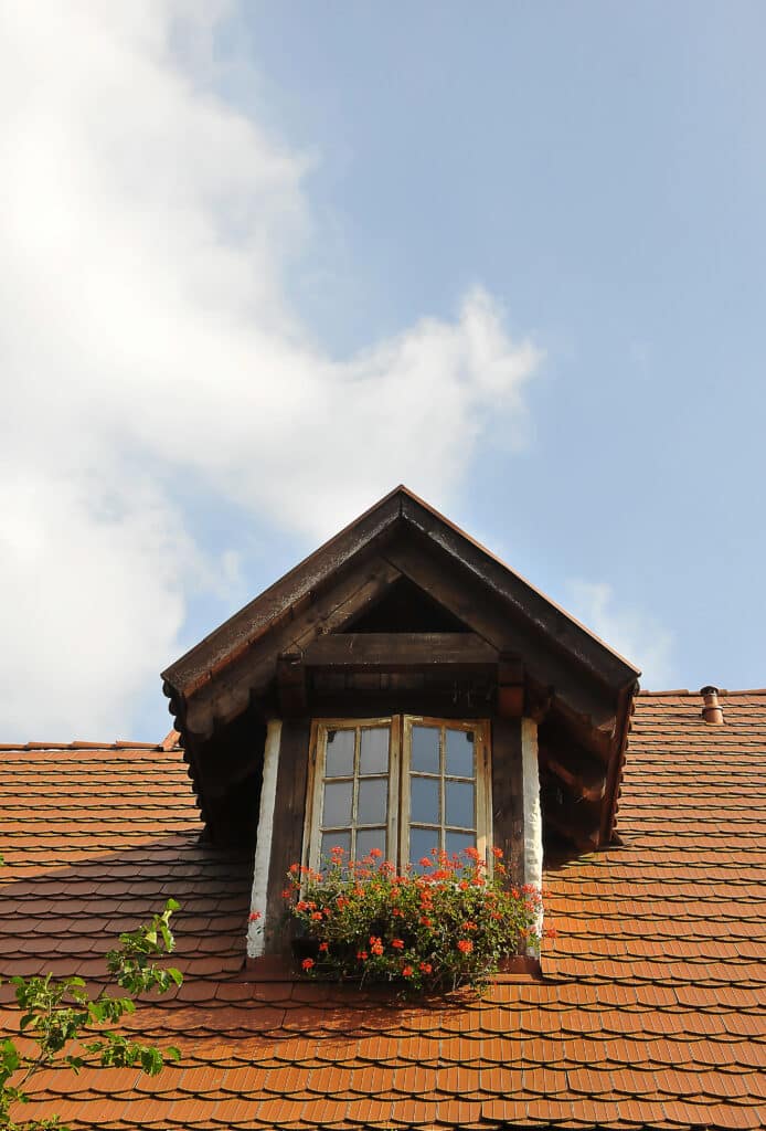 Rustic attic window with red flowers on terracotta roof under a blue sky.