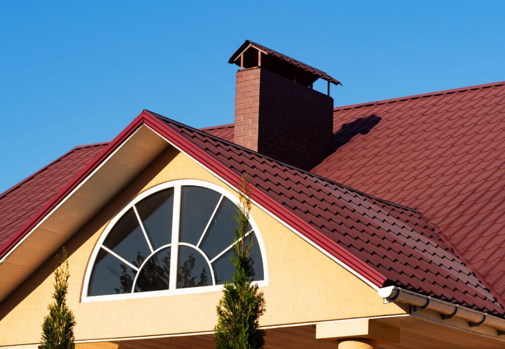 House with red metal roof, arched window, and chimney under clear blue sky.