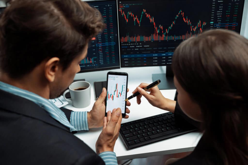 Professionals analyzing stock charts on phone and computer, focusing on market trends for financial strategy.