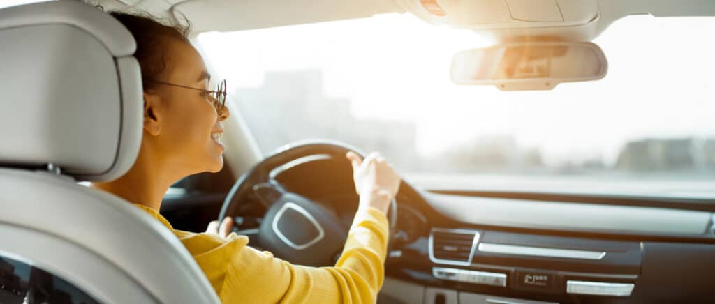Smiling woman driving car with sunlight streaming through windshield.