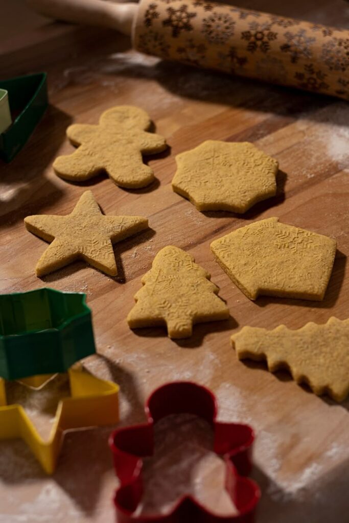 Christmas cookie dough cut into festive shapes on a floured table with a patterned rolling pin and cookie cutters.