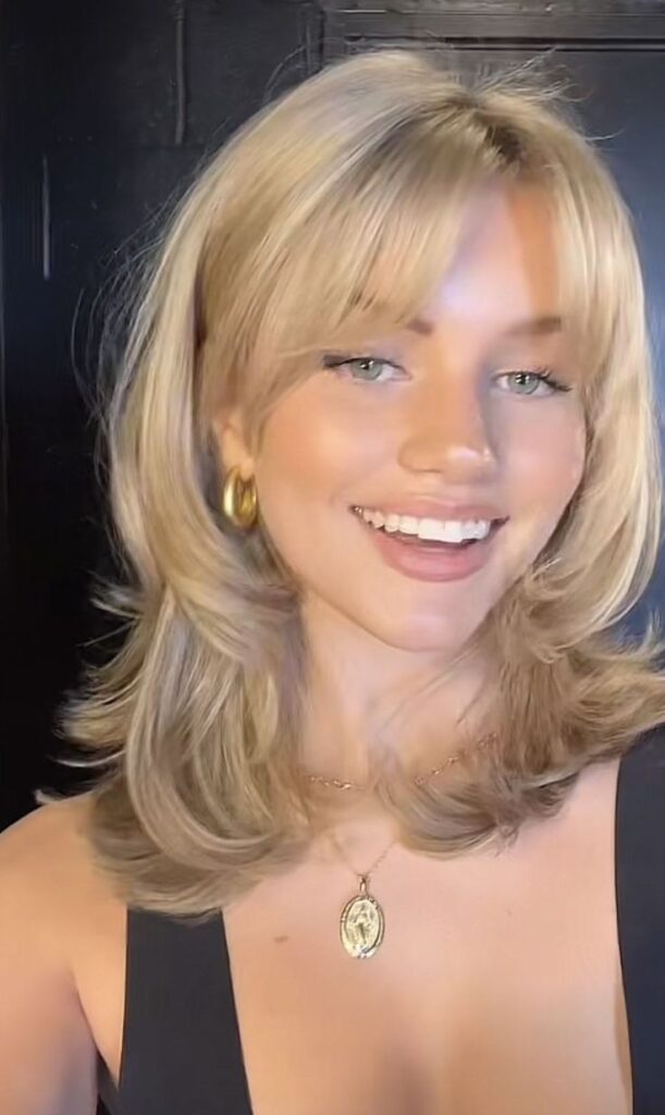 Smiling person with layered blonde hair and gold necklace, wearing gold hoop earrings against a dark background.