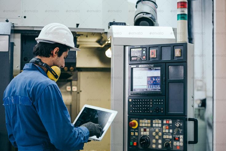 Factory worker operating CNC machine with tablet, wearing safety gear.