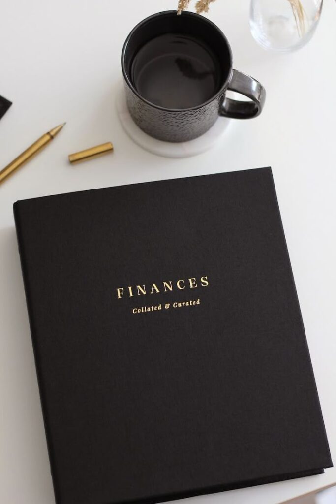 Black finance notebook, gold pen, and coffee on a white desk for an organized workspace aesthetic.