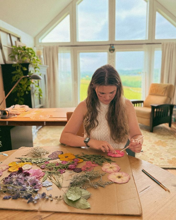 Every pressed flower frame we create is a true Woman crafting with dried flowers indoors, vibrant petals and leaves spread on table, large window in the background. | Sky Rye Design Woman crafting with dried flowers indoors, vibrant petals and leaves spread on table, large window in the background.