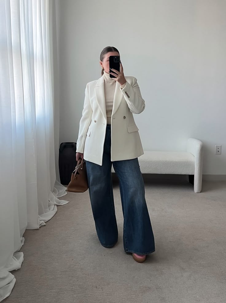 Woman in white blazer and jeans taking a mirror selfie in a stylish, minimalist room.