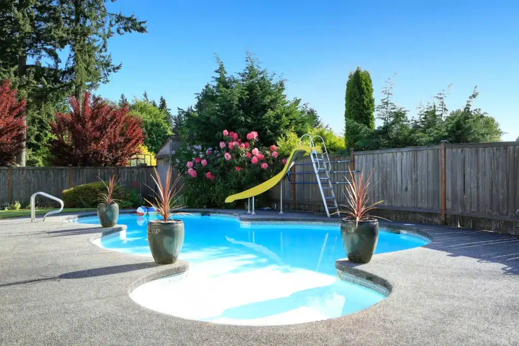 Fenced backyard with small beautiful swimming pool and playground shutterstock_447557716 Backyard pool with a yellow slide, potted plants, and clear blue sky. Perfect summer setting for relaxation. | Sky Rye Design Backyard pool with a yellow slide, potted plants, and clear blue sky. Perfect summer setting for relaxation.